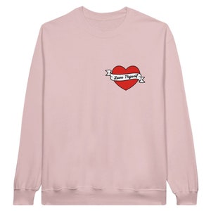 Puede incluir: Una sudadera rosa de manga larga con un corazón rojo y una pancarta que dice "Love Yourself" en la parte delantera.