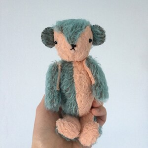 May include: A small, handmade plush toy bear with a teal blue and peach colored fur. The bear has black eyes and a black nose.