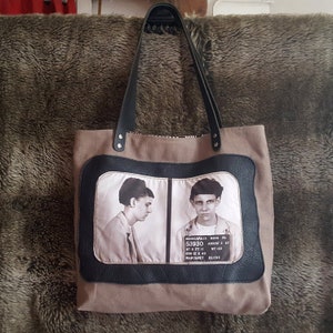 May include: A brown canvas tote bag with black leather handles and trim. The front of the bag features a black and white photo of two mugshots with the text "MINNEAPOLIS MINN PD 53930 AUGUST 3 67 HT & FT II DOS 12 2 45 MARGARET OLENE WT 133"