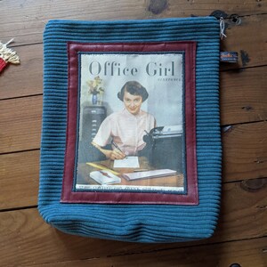 Peut inclure: Une pochette en velours c&ocirc;tel&eacute; bleu sarcelle avec une bordure rouge et blanche. La pochette pr&eacute;sente une couverture de magazine vintage avec le texte "Office Girl" et une photo d'une femme tapant sur un bureau.