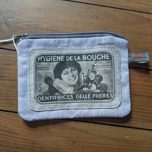Pode incluir: Uma bolsa de lona azul claro com zíper e acabamento prateado com um anúncio vintage de "Dentifrices Gelle Frères" com uma mulher e crianças. O texto do anúncio diz "Hygiène de la Bouche" e "Parfumeurs Paris".