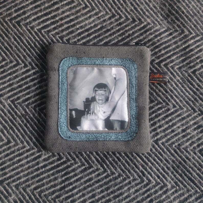 May include: A gray zippered pouch with a blue and white photo of a young girl blowing out candles on a birthday cake. The photo is framed with a blue border and the pouch has a small black tag with the text "Jackie" on it.