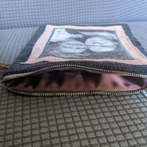 May include: A black zippered pouch with a pink fabric lining. The front of the pouch features a framed photo of two people.
