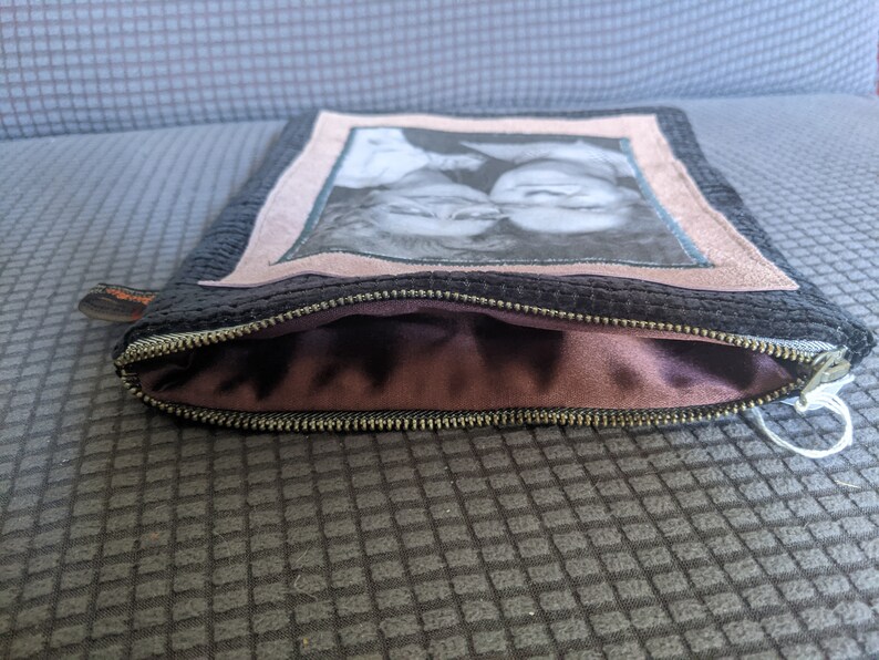 May include: A black zippered pouch with a pink fabric lining. The front of the pouch features a framed photo of two people.