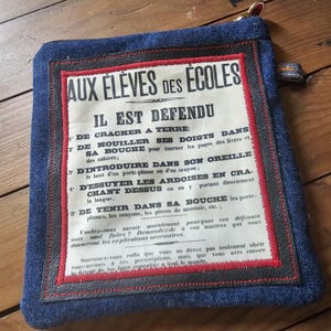 May include: A denim pouch with a vintage-style printed paper insert. The paper has French text in black, red, and white. The pouch has a red and black border and a zipper. The pouch is square and has a small tag.