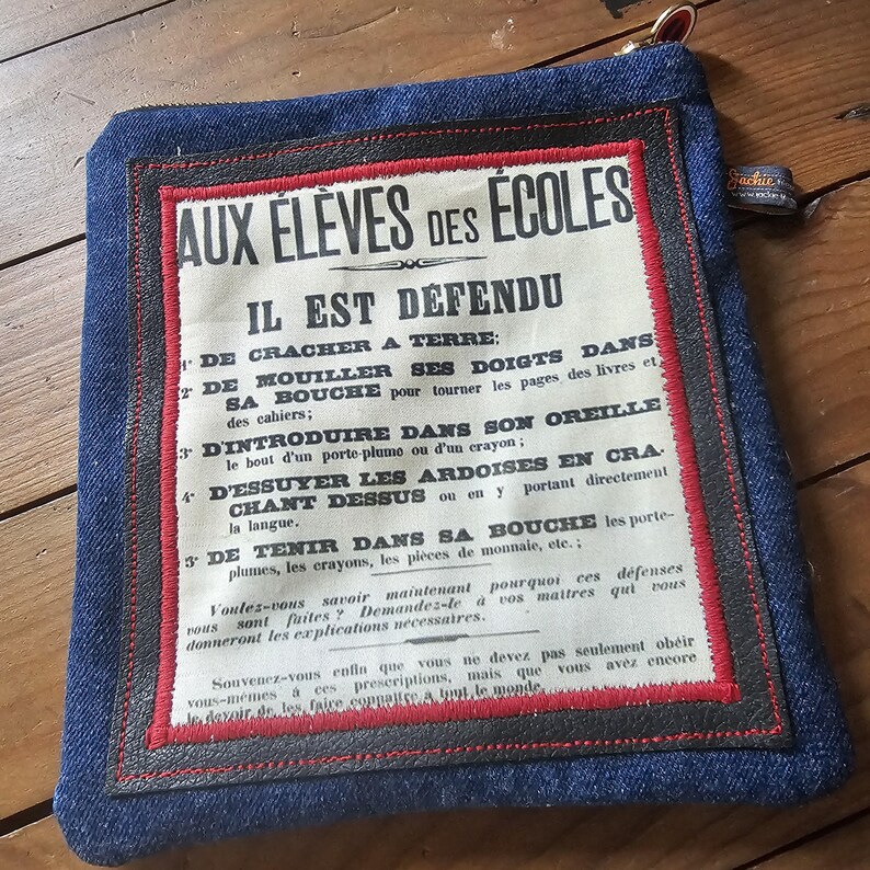 May include: A denim pouch with a vintage-style printed paper insert. The paper has French text in black, red, and white. The pouch has a red and black border and a zipper. The pouch is square and has a small tag.