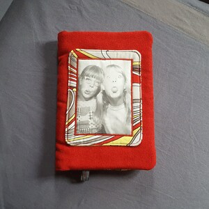 May include: Red fleece book cover with a photo of two girls with a yellow, red and brown patterned frame.