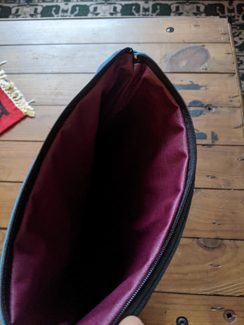 Peut inclure: Une pochette zipp&eacute;e turquoise avec une doublure bordeaux. La pochette est ouverte et la doublure est visible.
