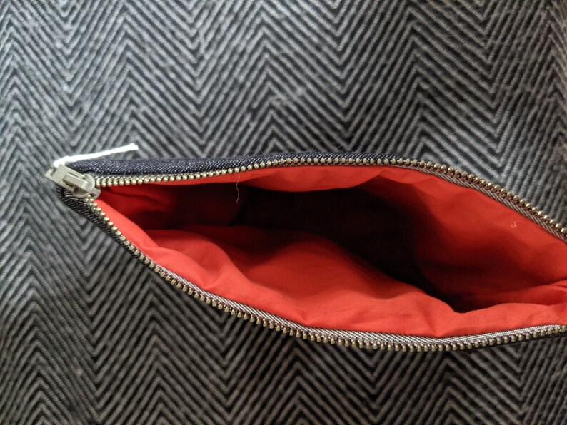 Puede incluir: Una bolsa de cremallera de denim negro con forro rojo. La bolsa est&aacute; abierta y la cremallera es visible.