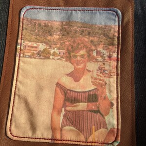 K&ouml;nnte beinhalten: Ein Vintage-Foto einer Frau mit Sonnenbrille und Badeanzug, die auf einem Strand mit einer bergigen Landschaft im Hintergrund steht. Das Foto ist in einem braunen Lederflicken mit roten N&auml;hten gerahmt.