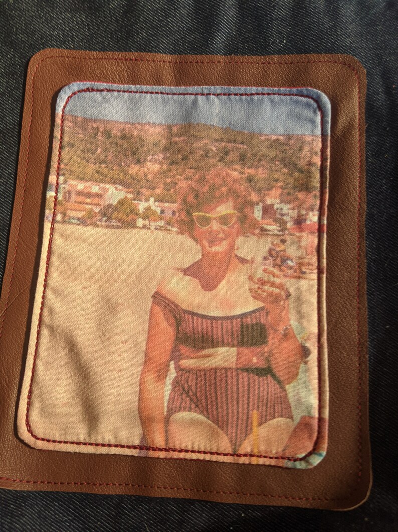 K&ouml;nnte beinhalten: Ein Vintage-Foto einer Frau mit Sonnenbrille und Badeanzug, die auf einem Strand mit einer bergigen Landschaft im Hintergrund steht. Das Foto ist in einem braunen Lederflicken mit roten N&auml;hten gerahmt.