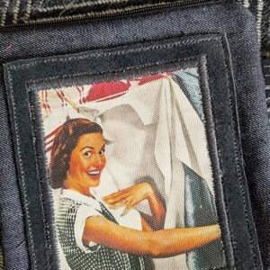 May include: A black fabric pouch with a zipper closure and a vintage-style patch featuring a woman smiling and holding a white cloth. The patch has a black border and is sewn onto the pouch.