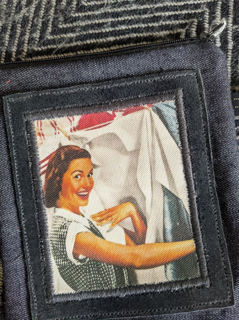 May include: A black fabric pouch with a zipper closure and a vintage-style patch featuring a woman smiling and holding a white cloth. The patch has a black border and is sewn onto the pouch.