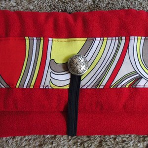 May include: A red felt clutch with a black strap and a silver button closure. The clutch features a colourful geometric pattern with yellow, white and brown accents.