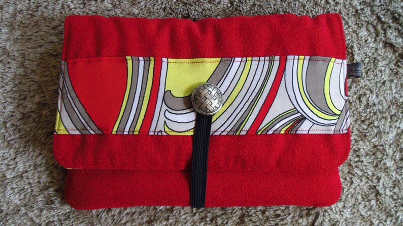 May include: A red felt clutch with a black strap and a silver button closure. The clutch features a colourful geometric pattern with yellow, white and brown accents.