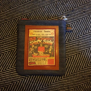 May include: A denim zipper pouch with a brown leather patch featuring a vintage poster for a "Shock Show" at the Paramount Theatre. The poster features a skeleton, a man in a red suit, and a woman in a black dress. The text on the poster reads "On Stage We Dare You To See... Dr. Brundell's Shock Show".