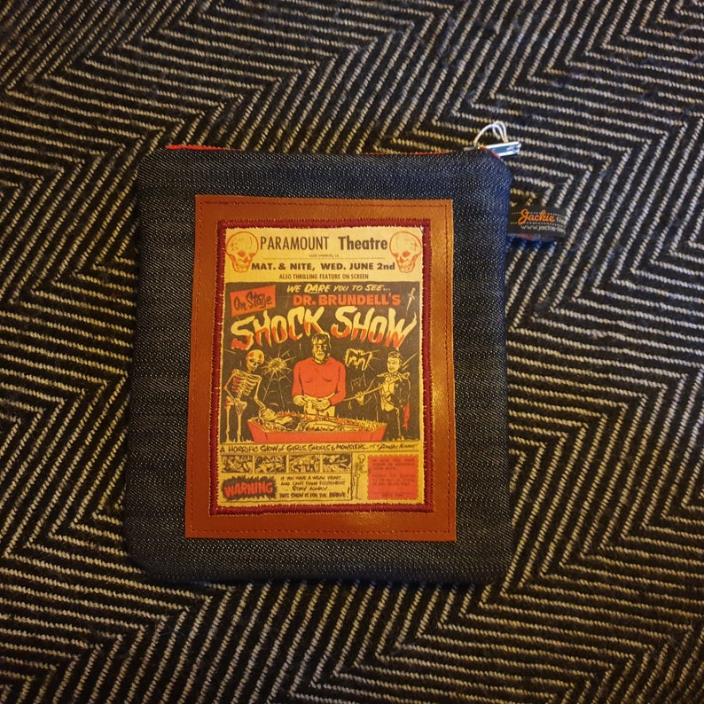 May include: A denim zipper pouch with a brown leather patch featuring a vintage poster for a "Shock Show" at the Paramount Theatre. The poster features a skeleton, a man in a red suit, and a woman in a black dress. The text on the poster reads "On Stage We Dare You To See... Dr. Brundell's Shock Show".