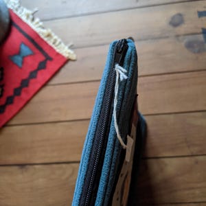 Peut inclure: Une pochette zipp&eacute;e bleu sarcelle avec une fermeture &eacute;clair noire et une &eacute;tiquette en ficelle blanche. La pochette est en tissu textur&eacute; et pr&eacute;sente un motif subtil.