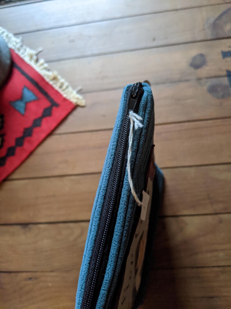Peut inclure: Une pochette zipp&eacute;e bleu sarcelle avec une fermeture &eacute;clair noire et une &eacute;tiquette en ficelle blanche. La pochette est en tissu textur&eacute; et pr&eacute;sente un motif subtil.