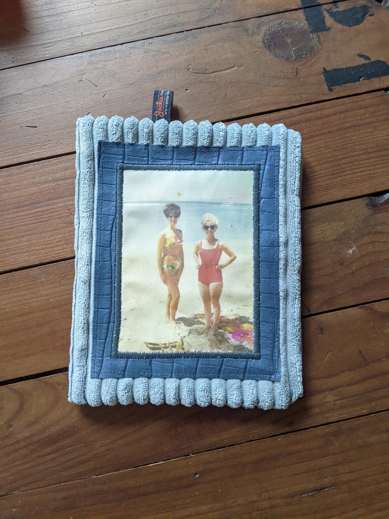 K&ouml;nnte beinhalten: Eine blaue Stofftasche mit einem Foto von zwei Frauen in Badeanz&uuml;gen am Strand. Das Foto ist mit einem blauen und braunen Rahmen eingefasst. Die Tasche hat ein kleines Etikett mit der Aufschrift "Sackit".