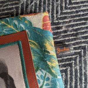 Pu&ograve; includere: Una cornice in pelle marrone con un bordo verde circonda una foto in bianco e nero. La cornice &egrave; fissata a uno sfondo in tessuto con un motivo a foglie tropicali in tonalit&agrave; di verde, giallo e rosso. Un'etichetta nera con testo bianco recita "Jackie Files" e "www.jackie-files.com".