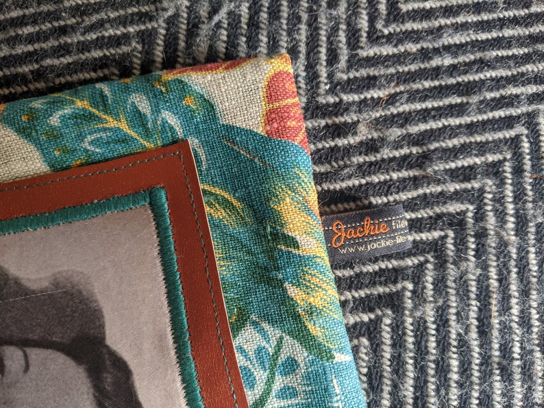 Pu&ograve; includere: Una cornice in pelle marrone con un bordo verde circonda una foto in bianco e nero. La cornice &egrave; fissata a uno sfondo in tessuto con un motivo a foglie tropicali in tonalit&agrave; di verde, giallo e rosso. Un'etichetta nera con testo bianco recita "Jackie Files" e "www.jackie-files.com".