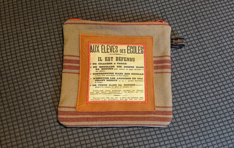 May include: A brown and tan striped fabric pouch with a zipper closure. The pouch has a leather patch with a vintage-style sign that reads "Aux Eleves des Ecoles Il Est Defendu" and lists a series of rules in French.