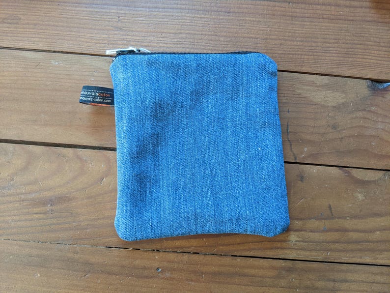 May include: A small blue denim zippered pouch with a black zipper pull and a black label with white text.