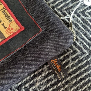 Puede incluir: Una bolsa de cremallera de denim negro con un parche de cuero negro y costuras rojas. El parche tiene un gr&aacute;fico de calavera y huesos cruzados y el texto "Theatre 2nd". La bolsa tiene una etiqueta de cuero marr&oacute;n con el texto "Jackie Filley" y el sitio web "www.jackie-filley.com".