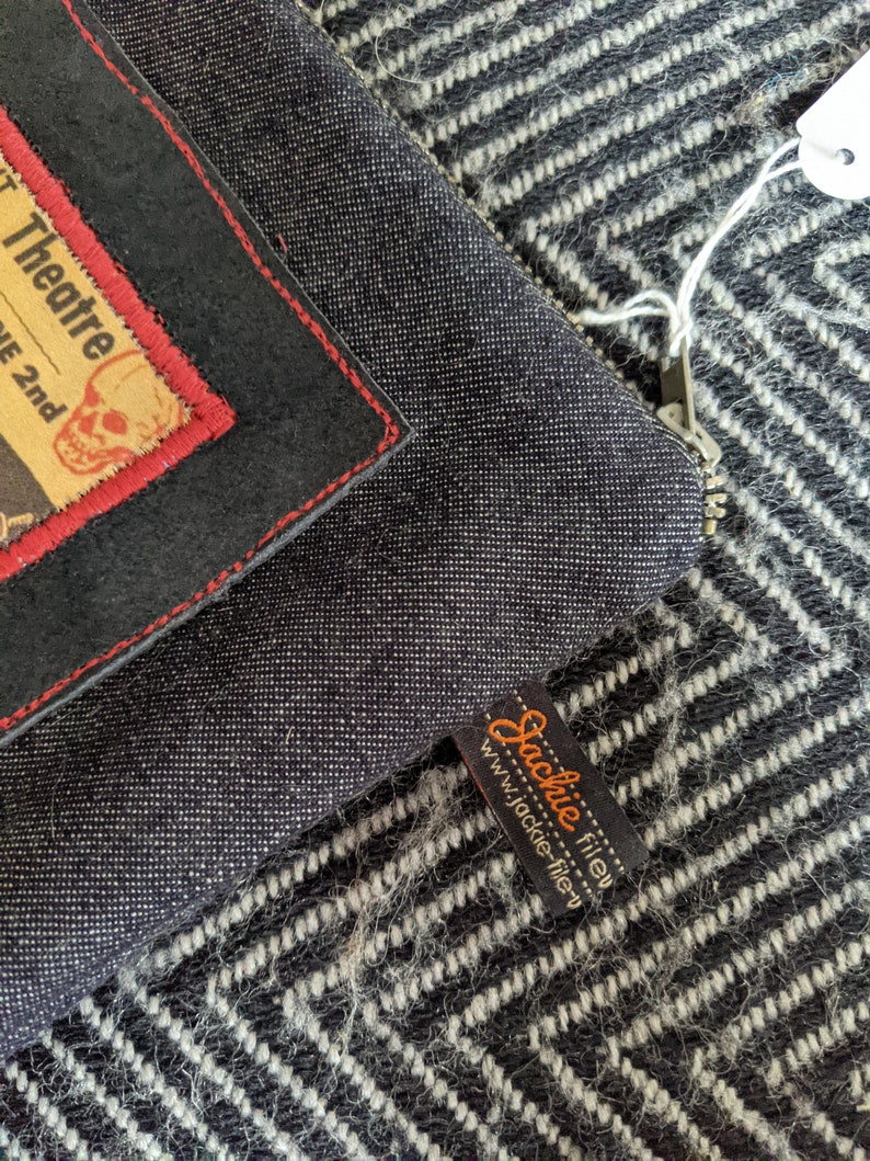 Puede incluir: Una bolsa de cremallera de denim negro con un parche de cuero negro y costuras rojas. El parche tiene un gr&aacute;fico de calavera y huesos cruzados y el texto "Theatre 2nd". La bolsa tiene una etiqueta de cuero marr&oacute;n con el texto "Jackie Filley" y el sitio web "www.jackie-filley.com".