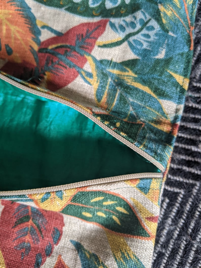 Pu&ograve; includere: Primo piano di una cerniera su una borsa in tessuto beige con una stampa a foglie tropicali in tonalit&agrave; di verde, giallo, rosso e blu.