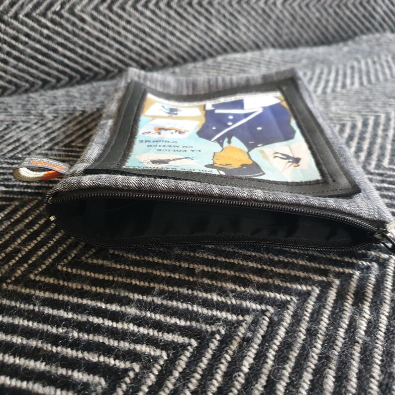 May include: A gray zippered pouch with a black zipper. The pouch has a rectangular patch with a vintage illustration of a police officer in uniform. The text on the patch reads "LA POLICE" and "POLICE NATIONALE".