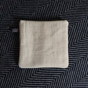 May include: A beige linen zippered pouch with a black zipper pull. The pouch is lying flat on a black and white patterned surface.