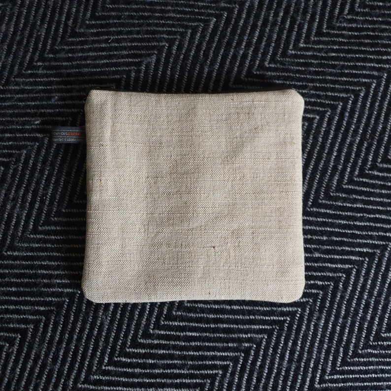 May include: A beige linen zippered pouch with a black zipper pull. The pouch is lying flat on a black and white patterned surface.