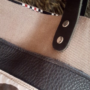 May include: Close-up of a beige canvas tote bag with a black leather strap and silver metal rivets. The strap has a black and white checkered trim.
