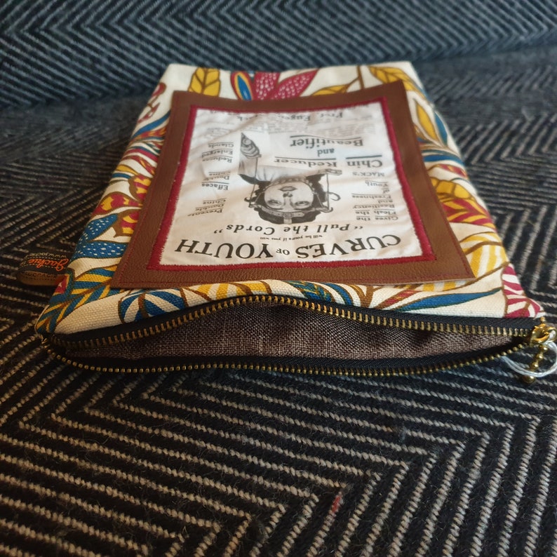 May include: A white zippered pouch with a brown leather patch. The patch features a vintage advertisement for a beauty product called "Chin Reducer and Beautifier." The text on the patch reads "CURVES OF YOUTH "Pull the Cords" Jacki Gares the Flesh the Castin MACK Chin Reducer and Beautifier Effacee Stande."