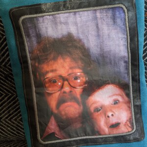 K&ouml;nnte beinhalten: Ein blaues Kissen mit einem schwarzen Rahmen und einem Foto von einem Mann und einem Kind. Der Mann tr&auml;gt eine Brille und einen Schnurrbart. Das Kind l&auml;chelt.