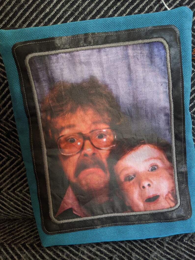 K&ouml;nnte beinhalten: Ein blaues Kissen mit einem schwarzen Rahmen und einem Foto von einem Mann und einem Kind. Der Mann tr&auml;gt eine Brille und einen Schnurrbart. Das Kind l&auml;chelt.