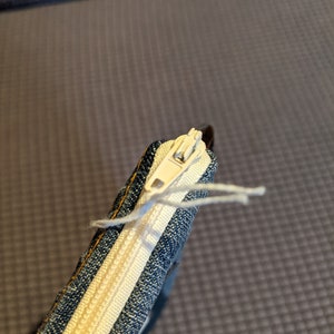 May include: A close-up of a denim zipper pouch with a white zipper and a white thread hanging from the zipper pull.
