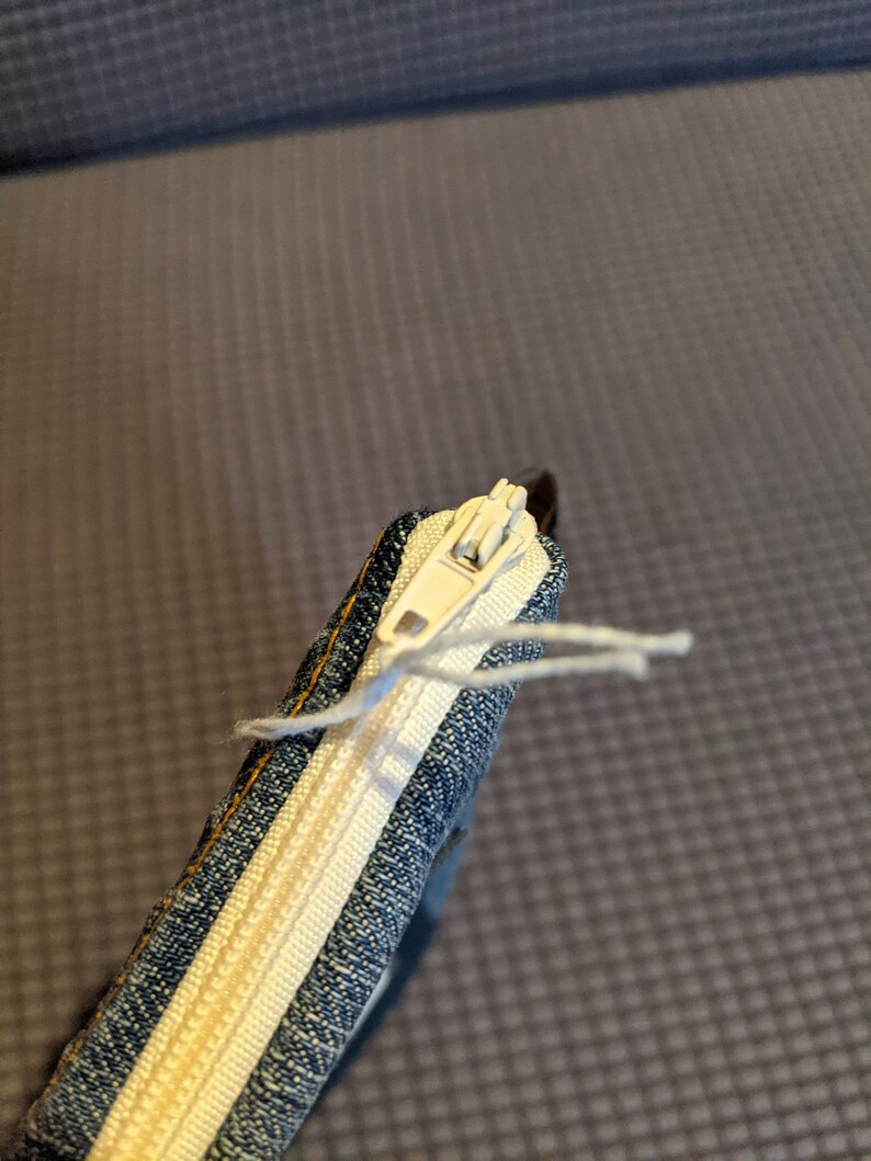 May include: A close-up of a denim zipper pouch with a white zipper and a white thread hanging from the zipper pull.