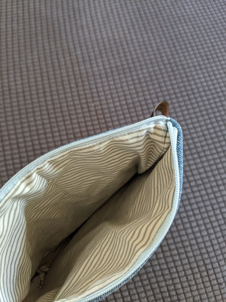 Puede incluir: Una bolsa de cremallera de mezclilla azul con un forro a rayas blanco y gris. La bolsa est&aacute; abierta y el forro es visible.