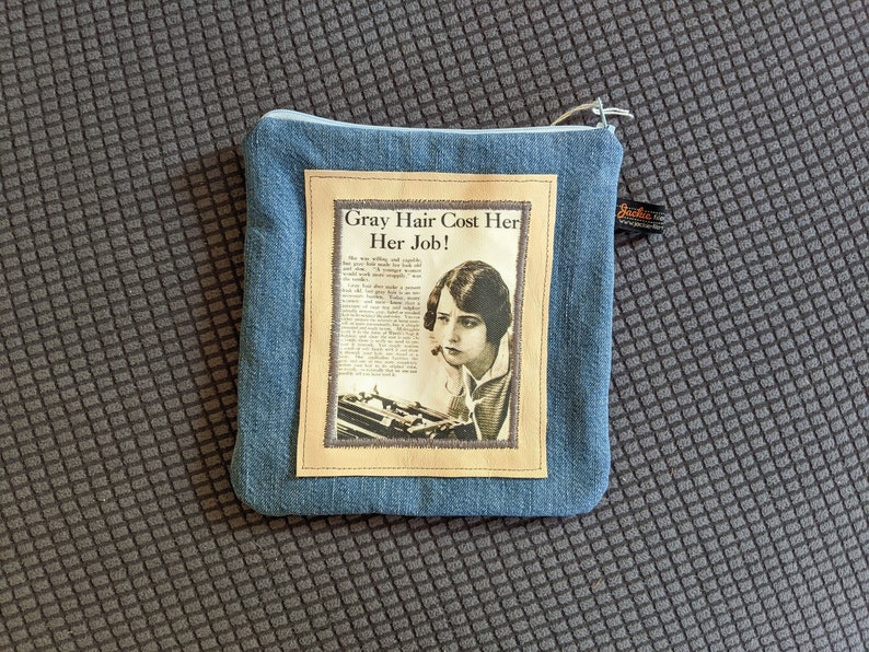 Puede incluir: Una bolsa de cremallera de mezclilla azul con un anuncio vintage de tinte para el cabello. El anuncio presenta a una mujer con una m&aacute;quina de escribir y el texto "Gray Hair Cost Her Her Job!"