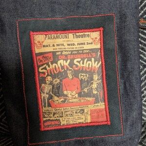 Puede incluir: Una bolsa de cremallera de denim negro con un parche bordado en rojo, blanco y negro que presenta un cartel de pel&iacute;cula vintage para "Dr. Brundell's Shock Show" en el Paramount Theatre. El cartel anuncia un "espect&aacute;culo horrible de chicas, demonios y monstruos" e incluye una advertencia para aquellos con corazones d&eacute;biles.