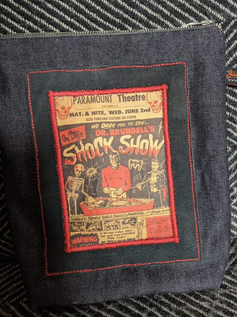 Puede incluir: Una bolsa de cremallera de denim negro con un parche bordado en rojo, blanco y negro que presenta un cartel de pel&iacute;cula vintage para "Dr. Brundell's Shock Show" en el Paramount Theatre. El cartel anuncia un "espect&aacute;culo horrible de chicas, demonios y monstruos" e incluye una advertencia para aquellos con corazones d&eacute;biles.