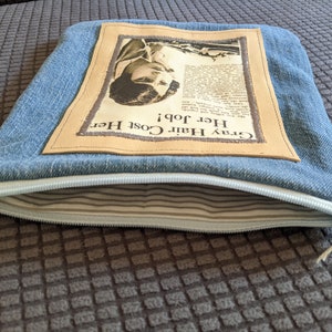 Puede incluir: Una bolsa de cremallera de mezclilla azul con un parche de cuero marr&oacute;n. El parche tiene un recorte de peri&oacute;dico vintage con el texto "Gray Hair Cost Her Her Job!"