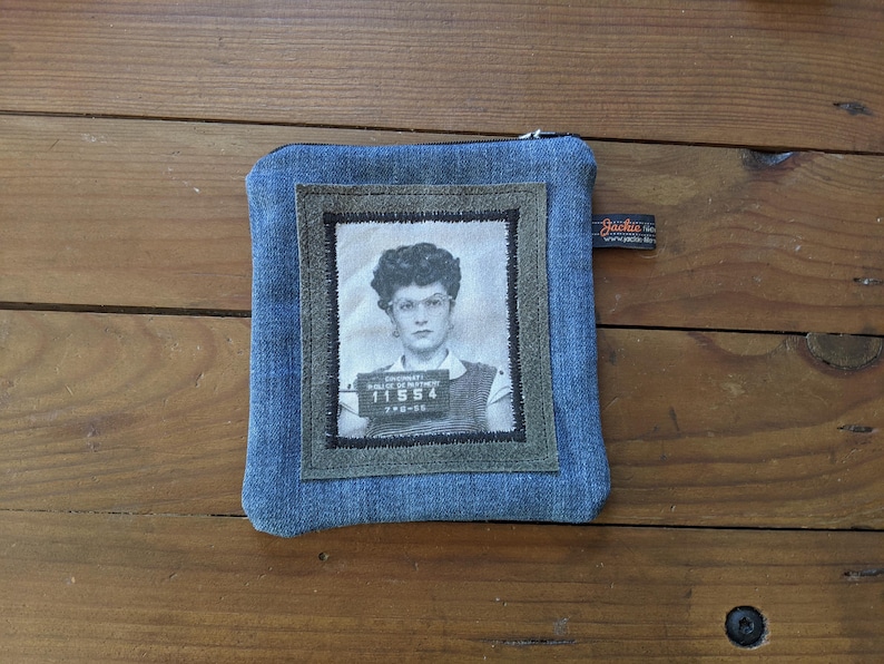 May include: A blue denim zippered pouch with a gray fabric patch featuring a black and white photo of a woman with a mugshot style photo ID. The ID reads "DEPARTMENT OF CORRECTIONS" and "11554".