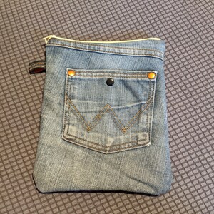 May include: A small blue denim pouch with a zipper closure and a front pocket. The pouch has two gold-coloured metal rivets on the top corners.