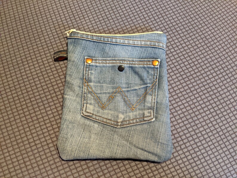 May include: A small blue denim pouch with a zipper closure and a front pocket. The pouch has two gold-coloured metal rivets on the top corners.