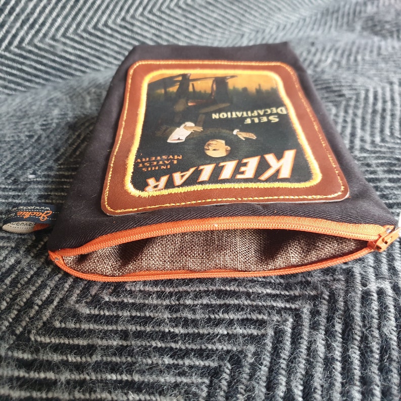 Pu&ograve; includere: Una pochette nera con cerniera e una toppa in pelle marrone con una pubblicit&agrave; vintage per "KELLAR, SELF DECAPITATION, IN THIS LATEST MYSTERY".