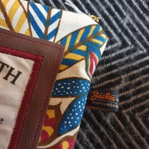 May include: A colorful fabric pouch with a brown leather trim and a label that says "Jackie" and "www.jackie.com". The pouch has a white background with blue, yellow, and red geometric patterns.
